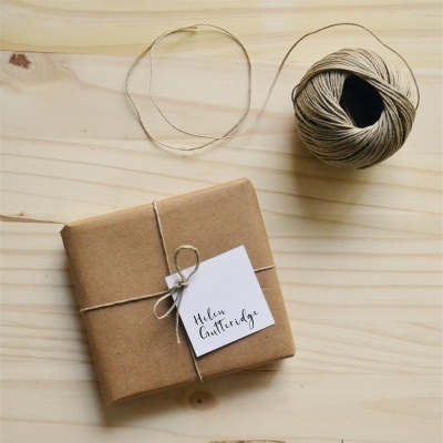 Pacote embrulhado em papel castanho com etiqueta e novelo de corda num fundo de madeira clara