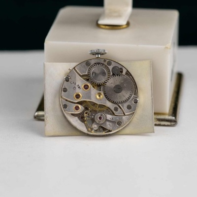 Mecanismo redondo de relógio em metal prateado com engrenagens visíveis sobre suporte branco