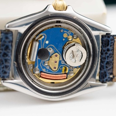 Relógio com mecanismo interno exposto, caixa prateada e bracelete de couro castanha com costura azul