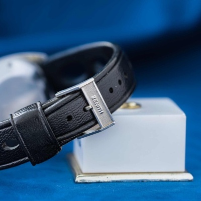 Relógio com bracelete preta em couro perfurado e fivela metálica com gravação 'NIXON', sobre fundo azul