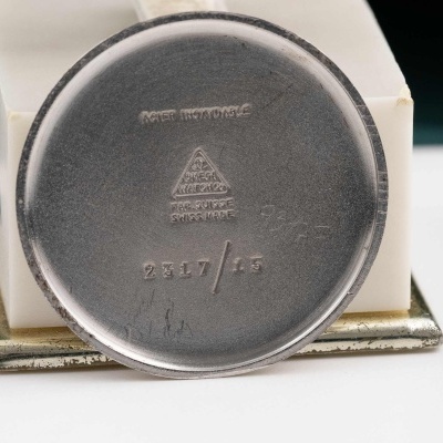 Relógio redondo de metal com gravações, sobre base branca e detalhes dourados.