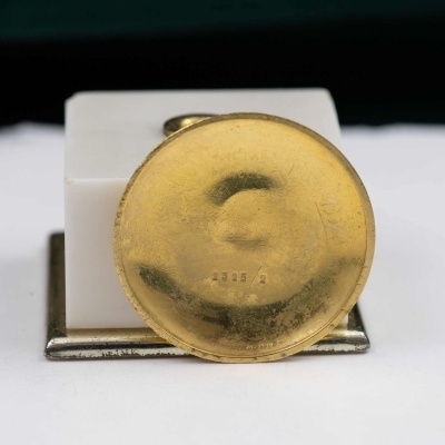 Objeto redondo dourado com número gravado sobre base branca e suporte metálico.