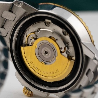 Relógio metálico aberto mostrando mecanismo interno e rotor dourado ETERNA-MATIC