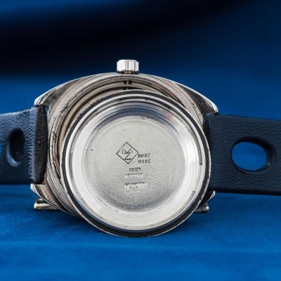 Relógio com caixa de metal, bracelete preta com orifícios, fundo da caixa com marcação, sobre superfície azul.