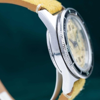 Relógio de pulso com caixa metálica prateada e bracelete amarela sobre fundo verde escuro