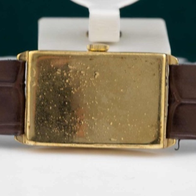 Relógio com caixa retangular dourada e pulseira castanha de couro com texto interno.