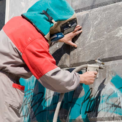 Pessoa com fato de proteção verde e vermelho a usar equipamento para jateamento de areia numa parede