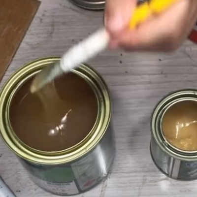 Duas latas abertas de tinta castanha e bege com pincel a aplicar tinta sobre mesa clara