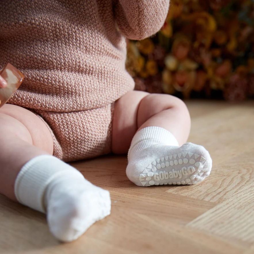 Meias brancas de bebé com sola antiderrapante GObabyGO, bebé sentado no chão com roupa de malha cor de rosa claro