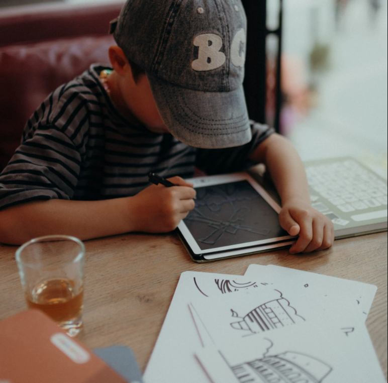 Criança a desenhar numa tablet digital com boné cinzento e t-shirt às riscas, mesa com desenhos em papel e chávena de vidro