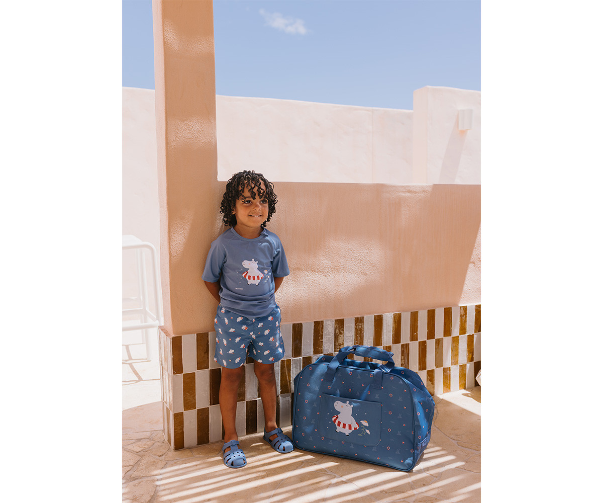 Criança e mala de viagem azul com estampa de hipopótamo encostadas numa parede bege com azulejos.
