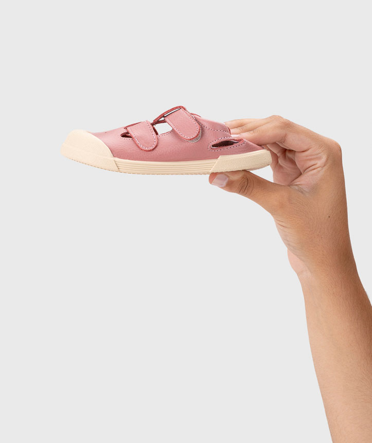 Sandália cor de rosa em pele com sola branca e duas tiras com velcro, segurada por mão contra fundo branco