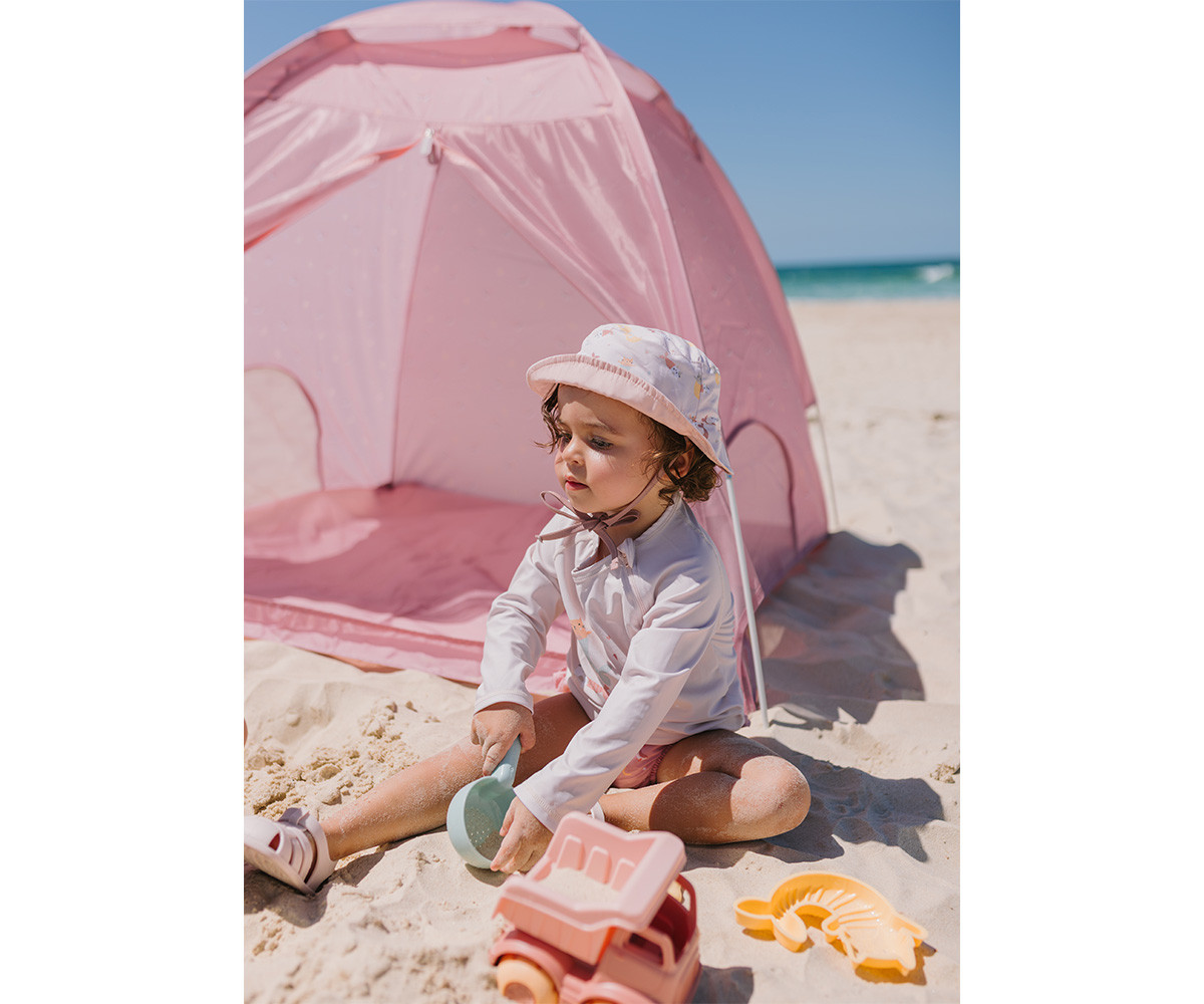 Criança sentada na areia com chapéu branco e tenda rosa atrás