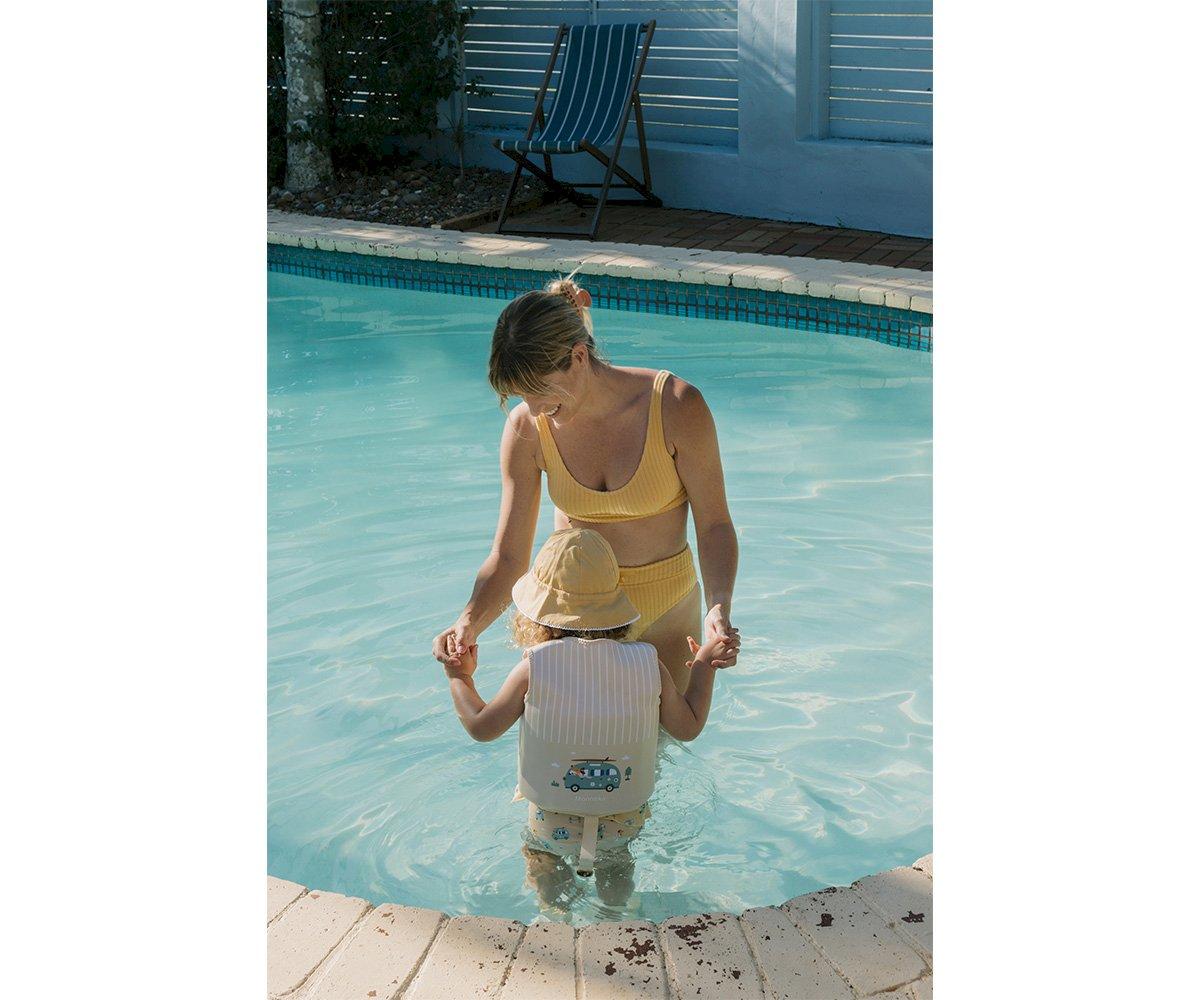 Mulher e criança na margem de piscina, criança com colete salva-vidas e chapéu