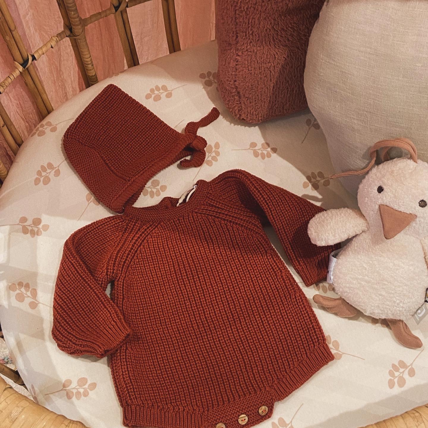 Roupa de bebé em malha castanha, camisola com botões e gorro, sobre almofada creme com padrão castanho, junto a peluche de pato.