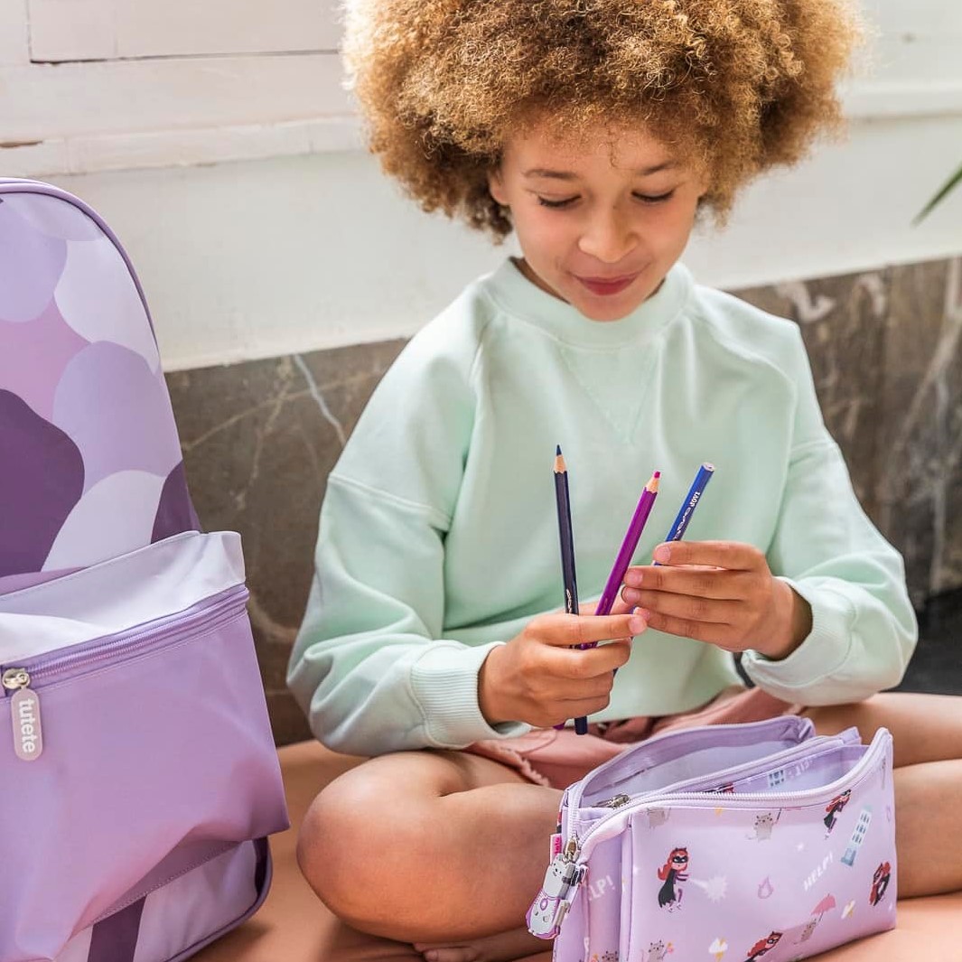 Criança com mochila e estojo lilás segurando lápis de cor, em ambiente interior