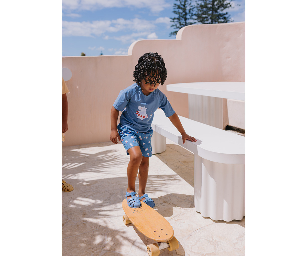 Criança de roupa azul a andar de skate de madeira num espaço exterior com mesa e bancos brancos