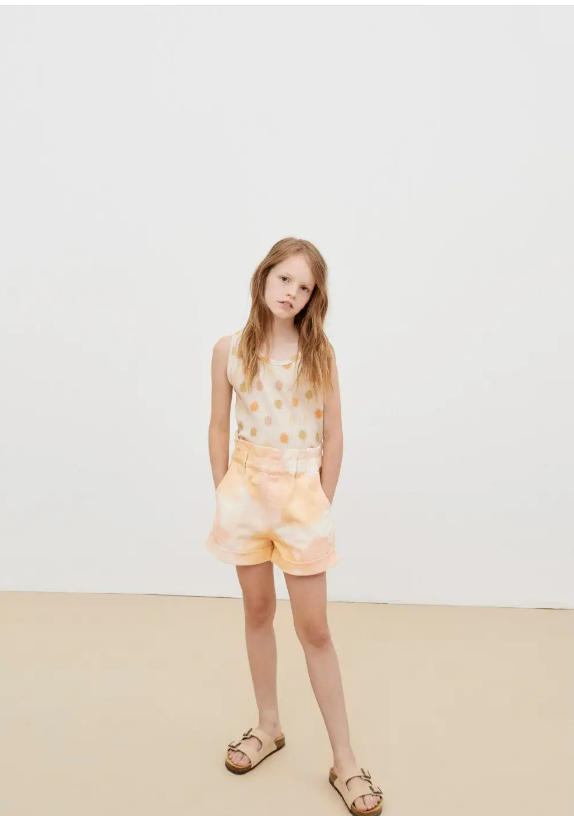 Menina com top branco com bolinhas coloridas, calções tie-dye cor de pêssego e sandálias bege, em fundo branco e chão bege.