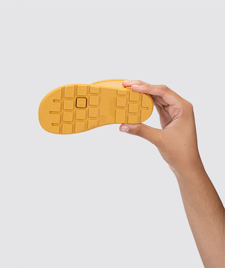 Sandália infantil amarela vista pelo solado, mão a segurá-la, fundo cinzento claro