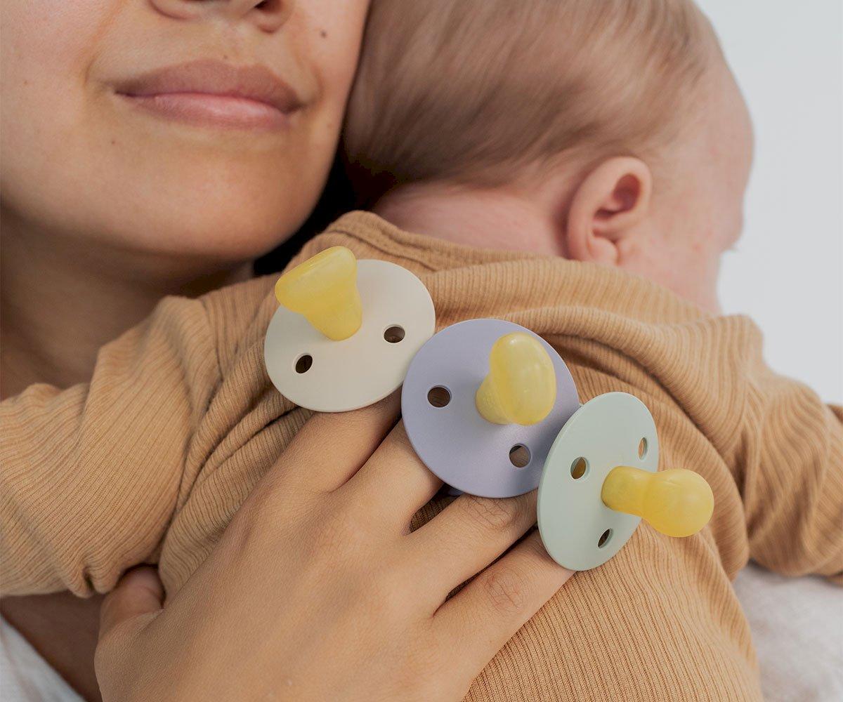 Três chupetas coloridas numa mão segurando um bebé com roupa castanha clara