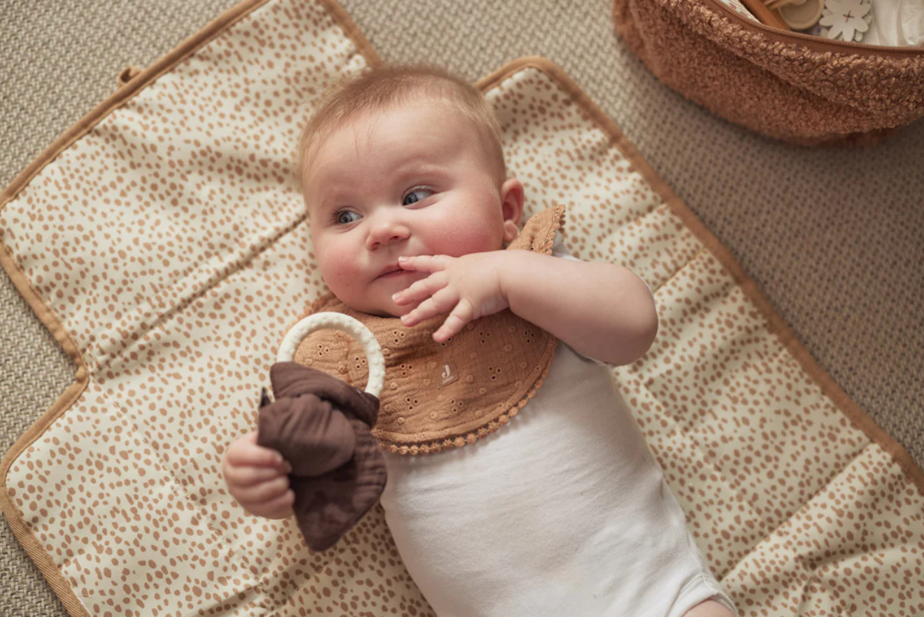 Bebé deitado com babete castanho segurando brinquedo de tecido castanho em colchão creme com padrão castanho