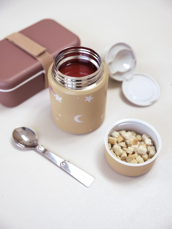 Termo bege decorado com estrelas e lua, líquido vermelho, recipiente com cubos de pão, colher metálica e caixa rosa