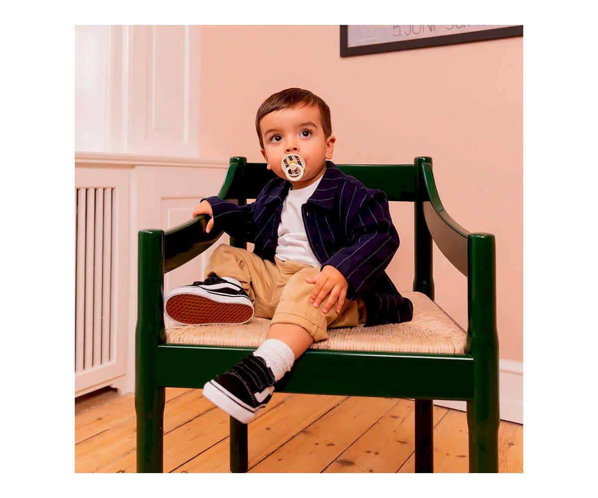 Criança sentada numa cadeira verde com assento em palhinha, roupa casual e chupeta na boca