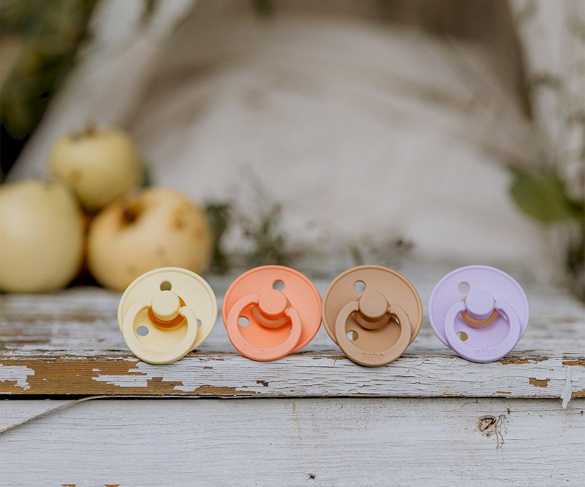 quatro chupetas coloridas em cores pastel alinhadas numa superfície rústica com maçãs ao fundo