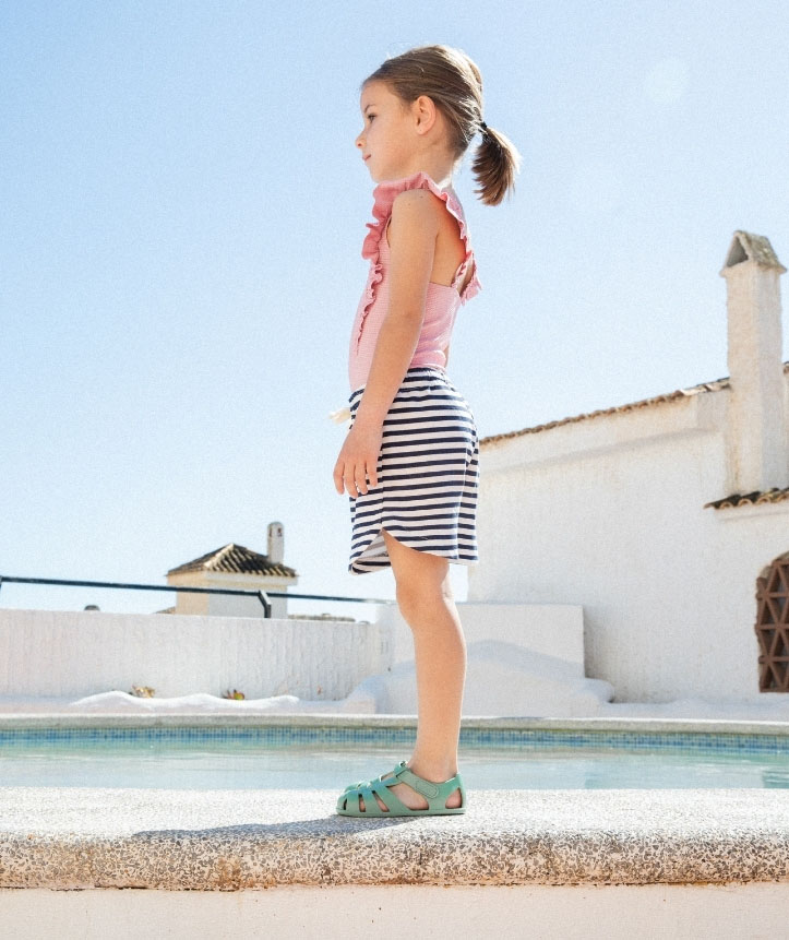 Criança com roupa rosa, saia riscada e sandálias verdes junto a piscina ao ar livre