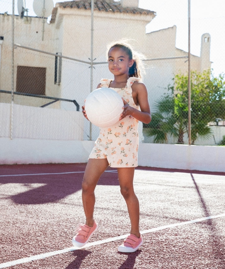 Menina em campo de desporto com macacão floral bege e sapatos rosa segurando bola de voleibol branca