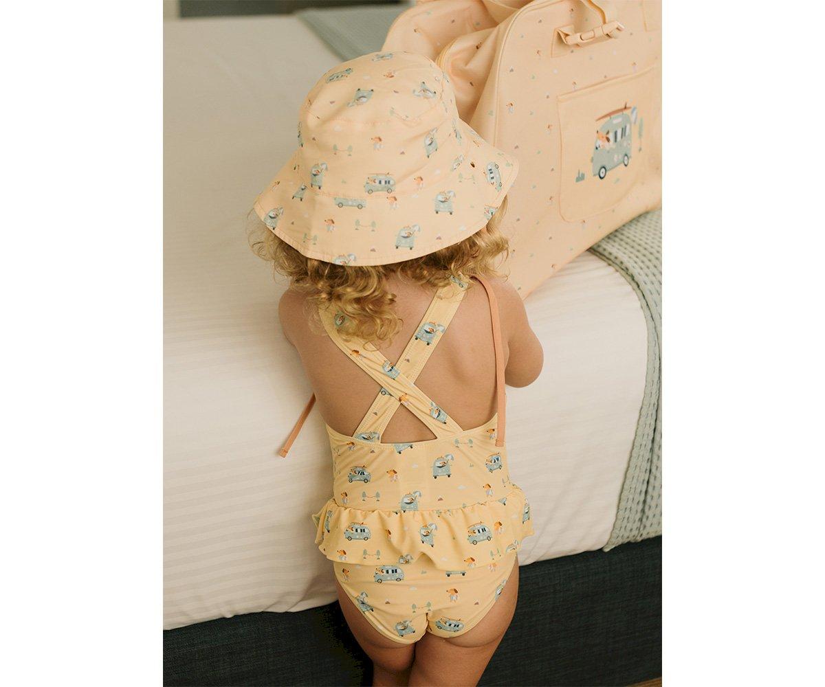 Criança de costas usando fato de banho amarelo e chapéu de sol no interior, próximo a cama e saco de viagem bege.