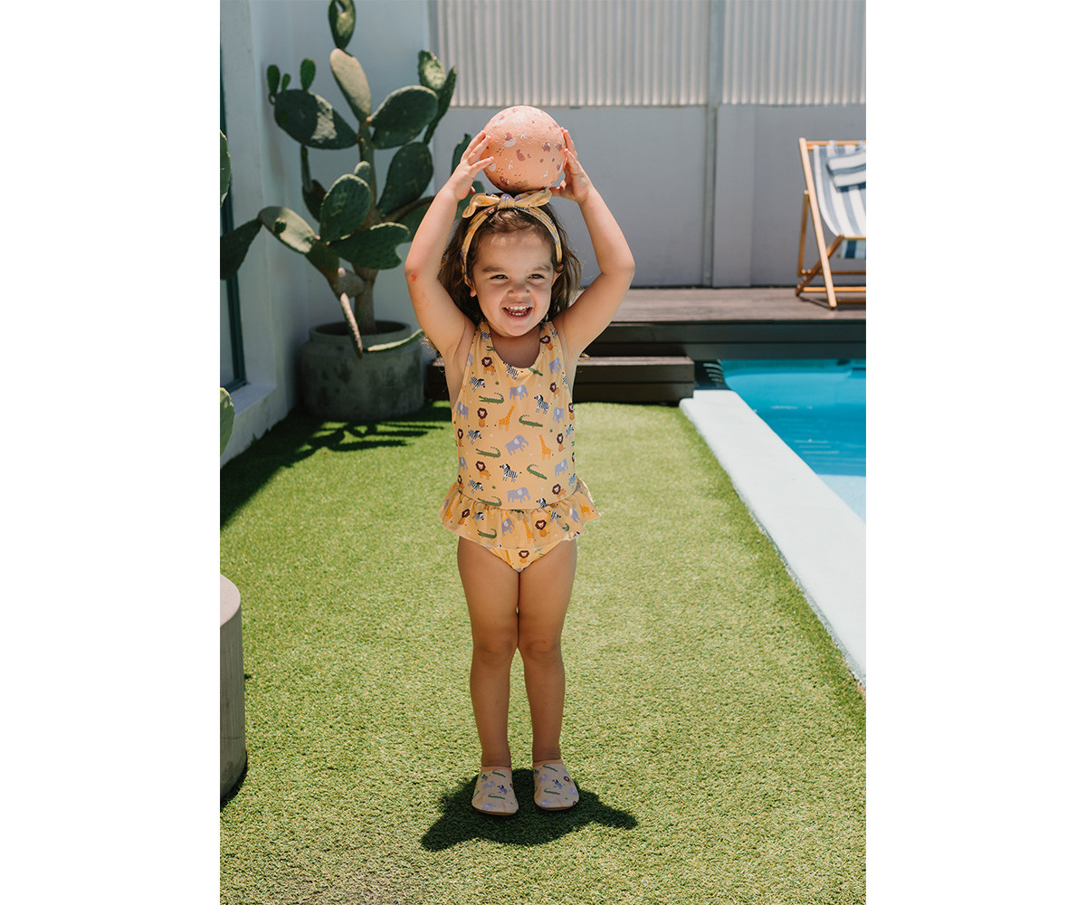 Criança a sorrir com fato de banho amarelo com animais, segurando bola cor-de-rosa junto a piscina