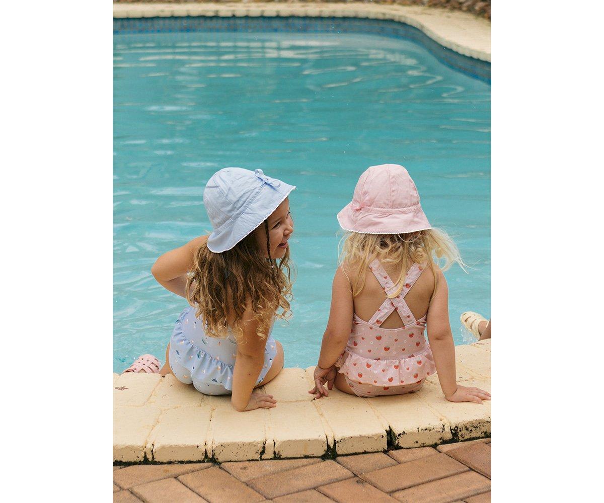 Duas crianças sentadas à beira de piscina com fatos de banho e chapéus coloridos
