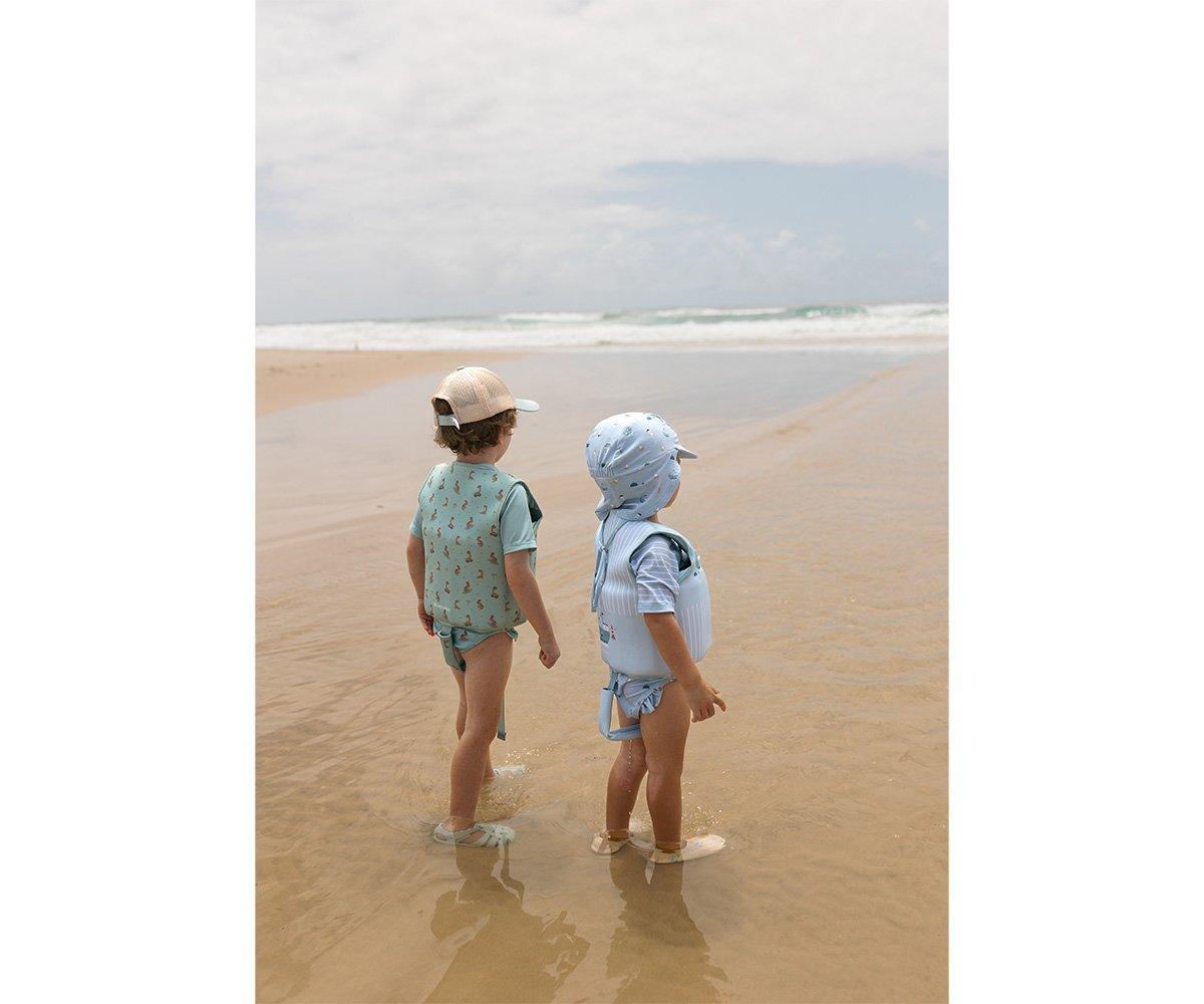 Dois bebés na praia com roupa de banho e chapéus coloridos de pé na areia molhada