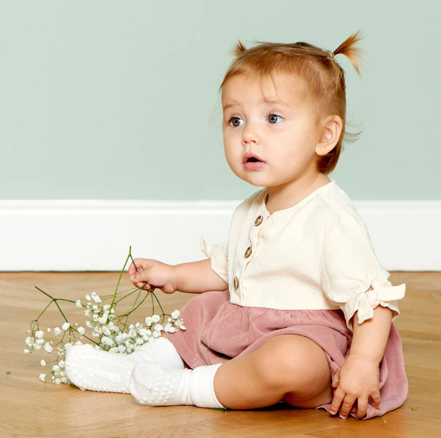 Bebé com vestido bege e saia rosa clara sentada no chão segurando flores brancas