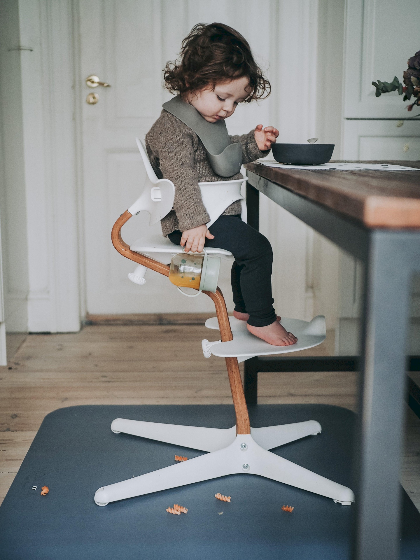 Criança sentada em cadeira alta para refeições com suportes para pés, próxima a mesa