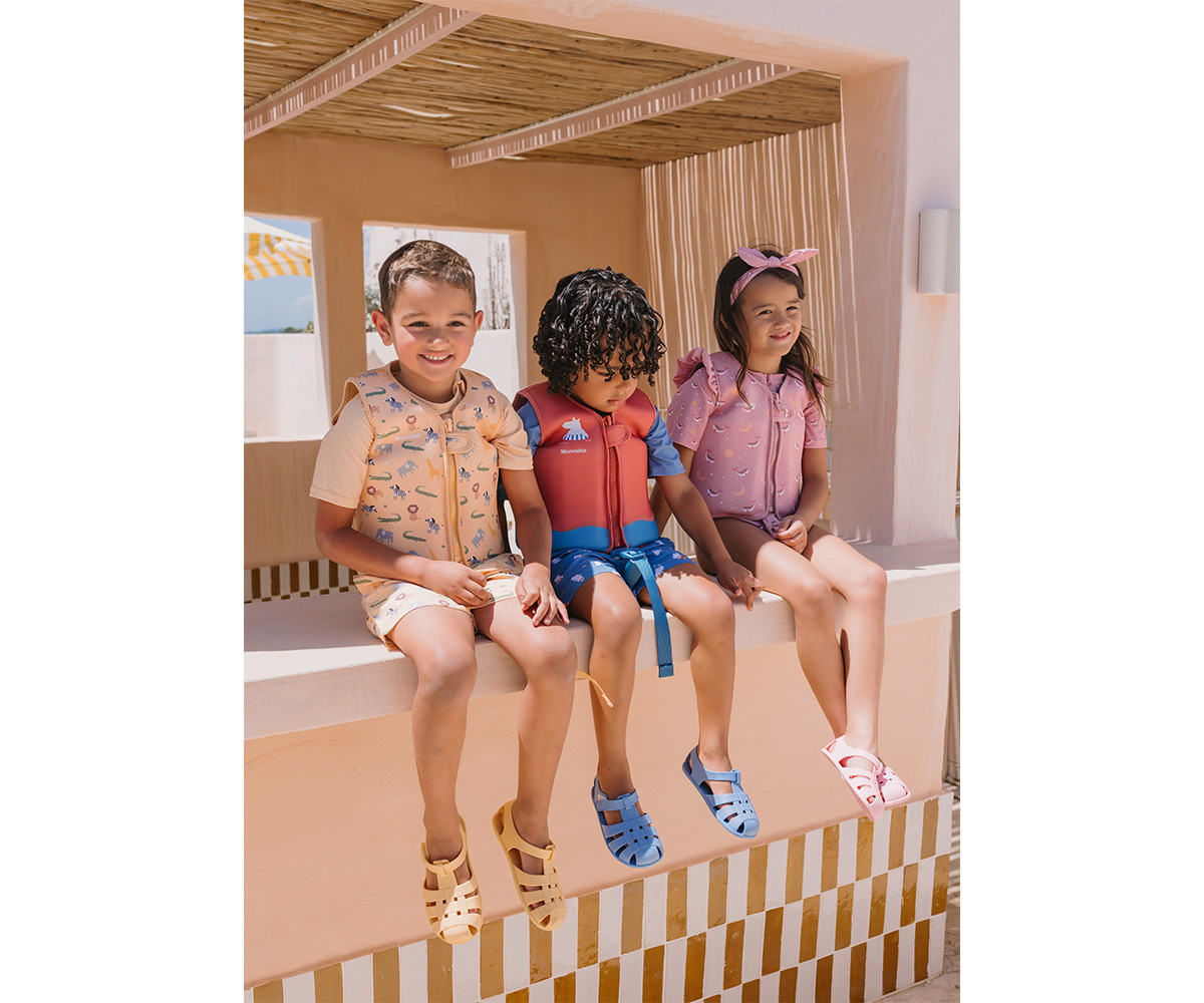 Três crianças com coletes salva-vidas coloridos e sandálias sentadas em muro exterior.