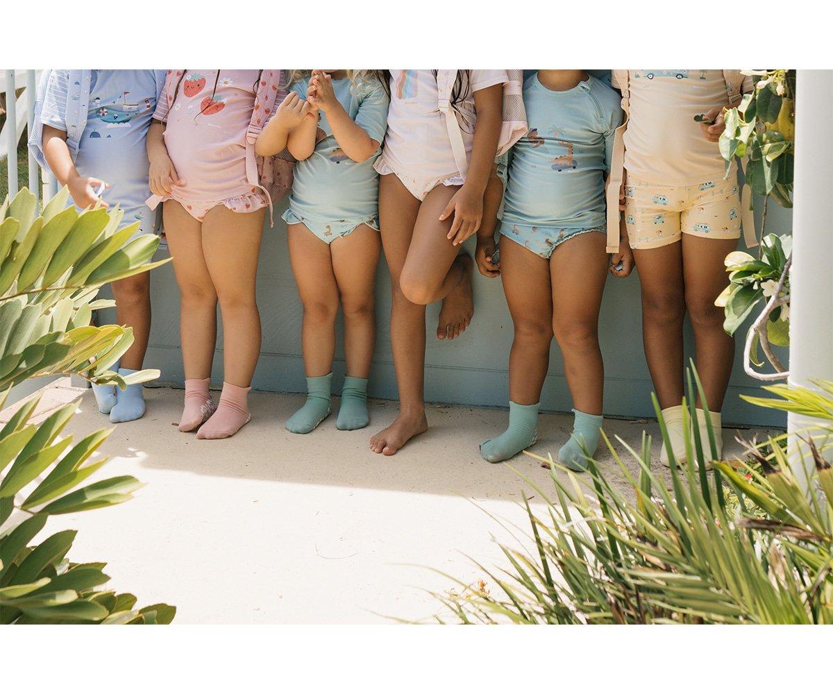 Quatro crianças em bodies coloridos com padrões, meias pastel, numa varanda com plantas verdes.