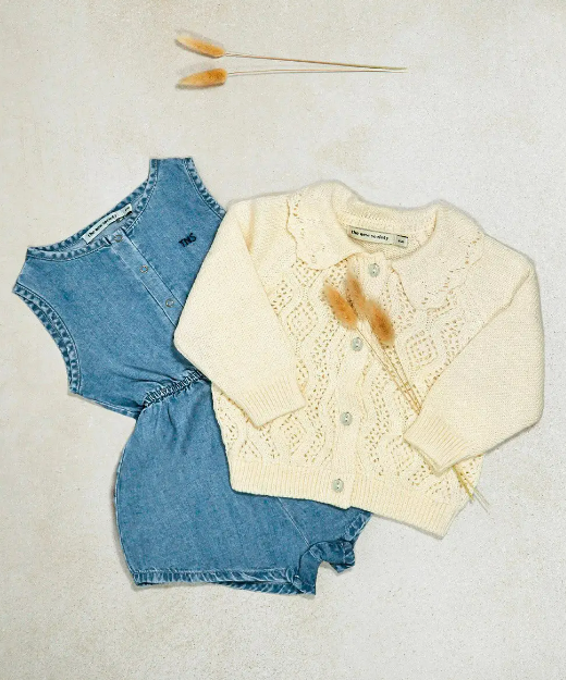 Conjunto de roupa de bebé em ganga azul e casaco creme de malha com botões, em fundo claro com flores secas