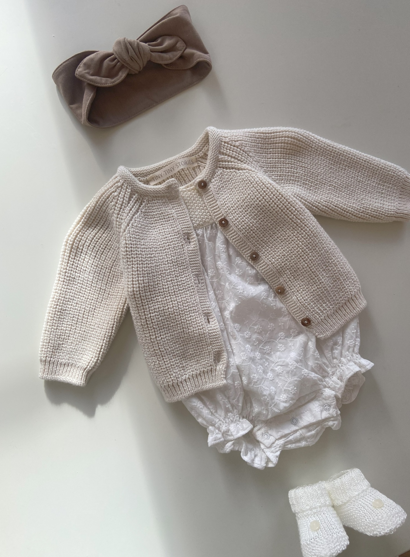 Conjunto de bebe com cardigan bege, body branco floral, botas brancas e faixa de cabelo castanha.