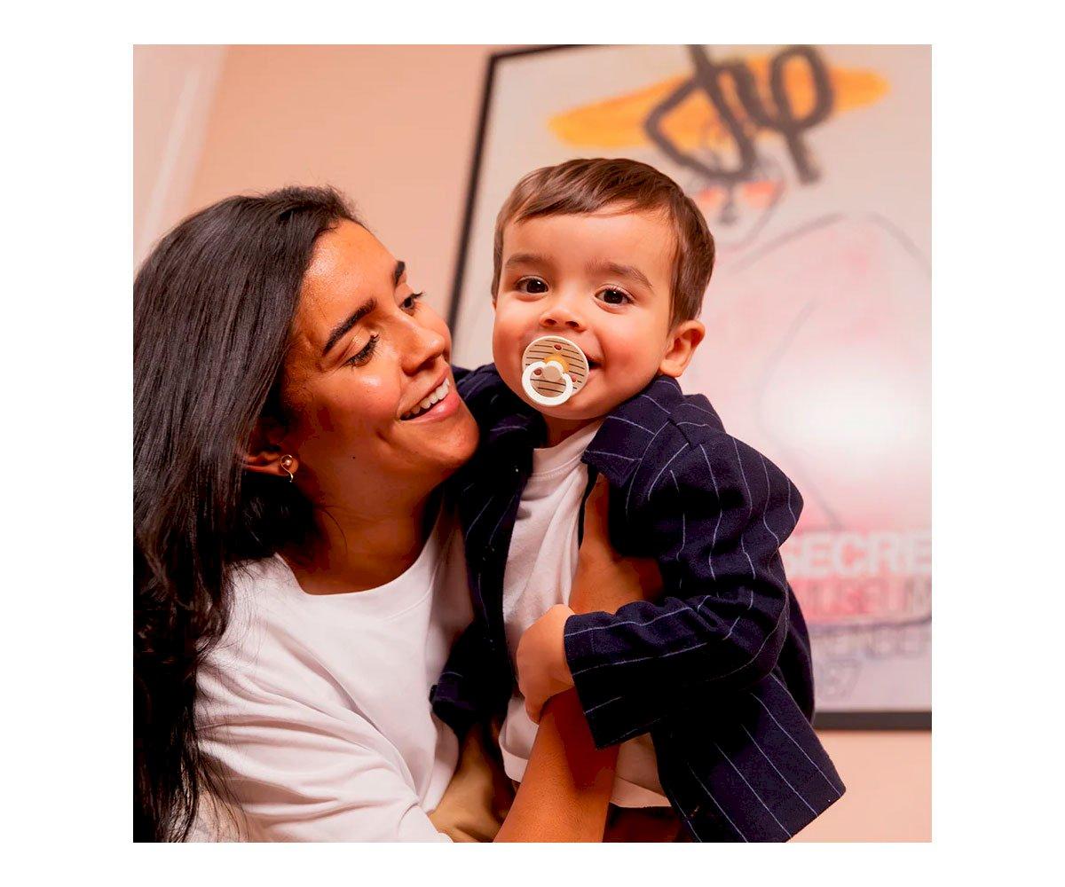 Mulher segura criança com blusão às riscas e chupeta em fundo com quadro