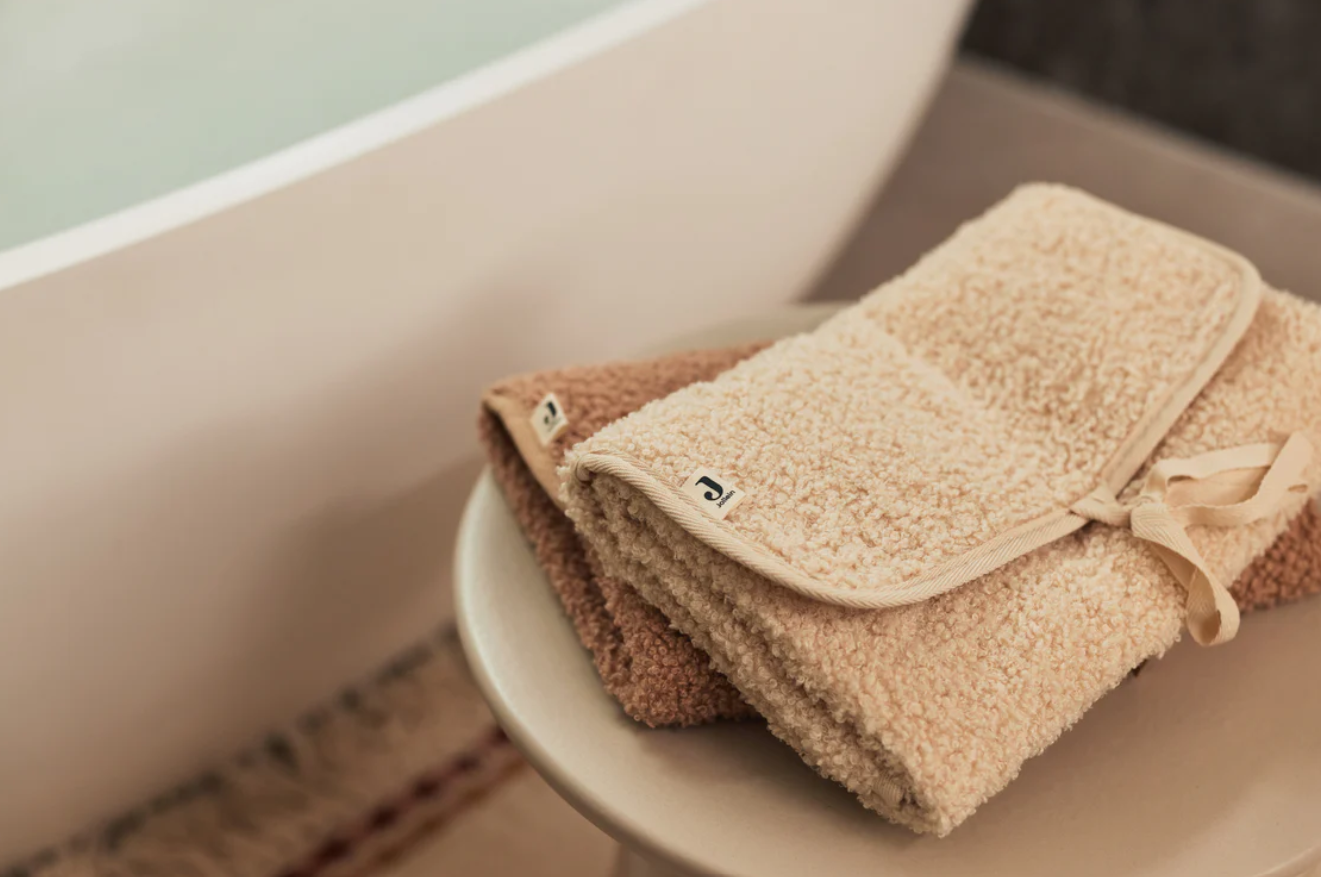 Toalhas felpudas bege e castanho claro dobradas numa superfície redonda branca perto de uma banheira