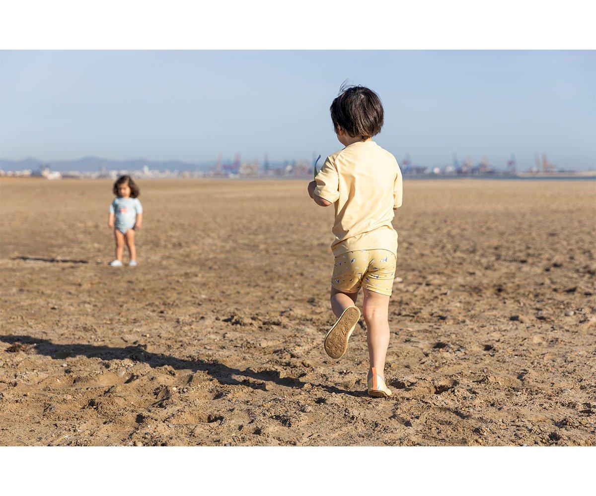 Duas crianças na praia, uma de costas com roupa clara e outra ao fundo com fato de banho azul-claro