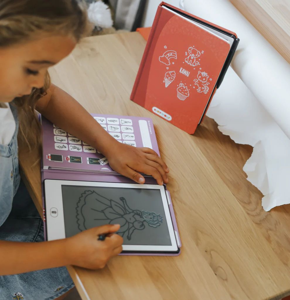 Menina a desenhar numa tablet com capa lilás e caderno laranja KAWAI numa mesa de madeira.