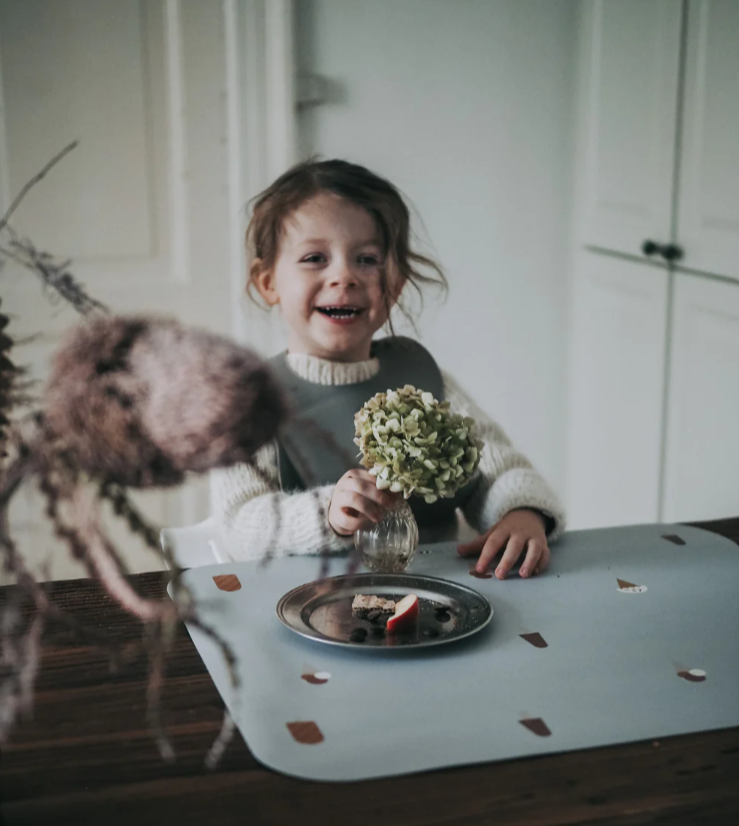 Criança sorridente sentada à mesa com flor em vaso e prato metálico