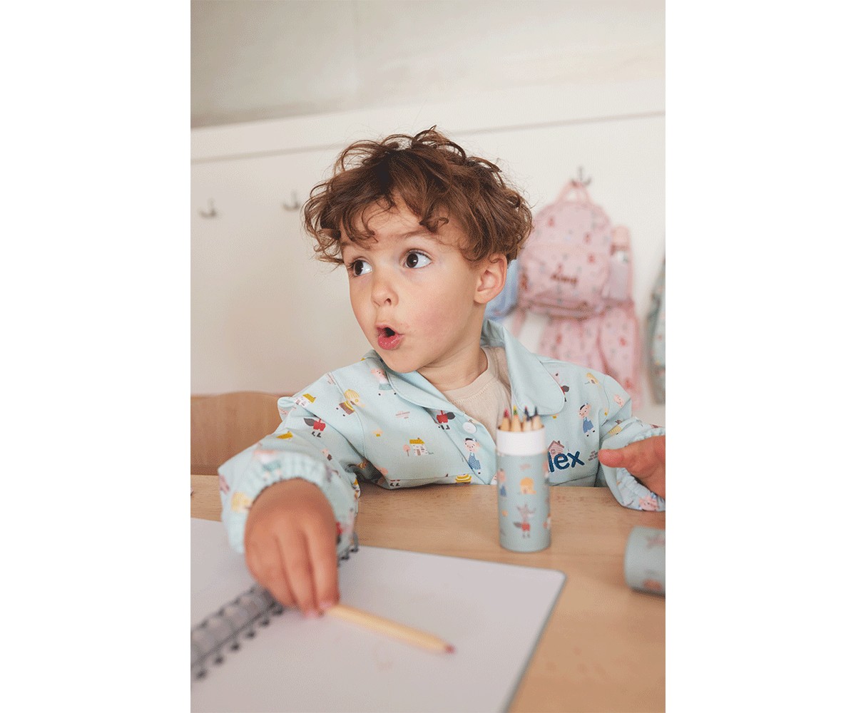 Criança com pijama azul claro desenhando com lápis num caderno numa mesa de madeira clara