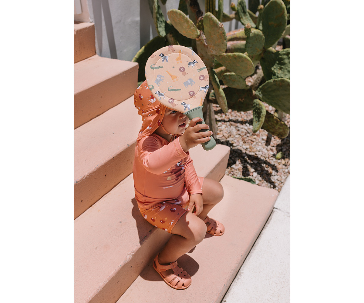 Criança com roupa de banho e chapéu cor de pêssego sentada em escadas, segurando raquete estampada, junto a cactos.