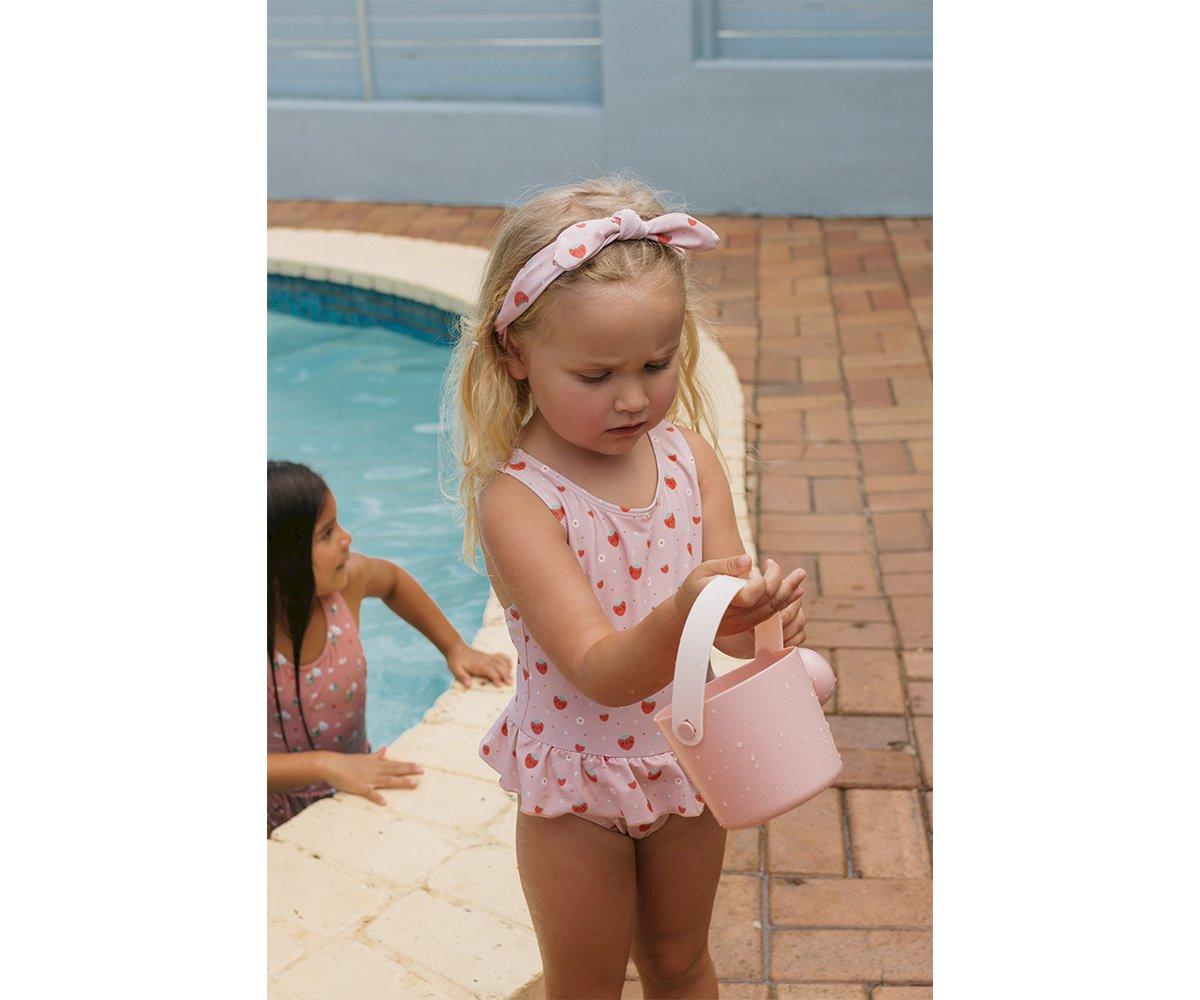 Menina pequena com maiô rosa de corações vermelhos e faixa no cabelo junto a piscina segurando cesta cor de rosa