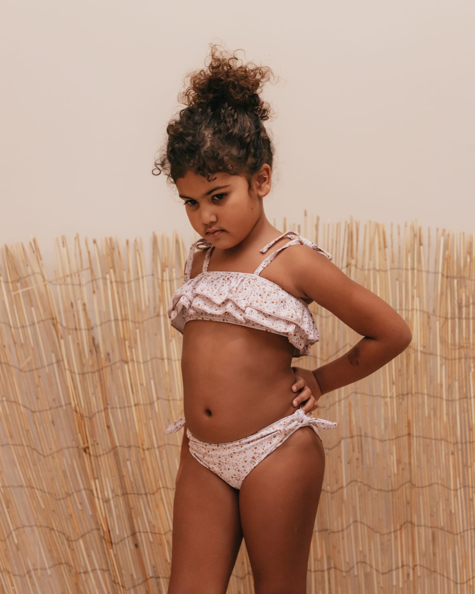 Menina de biquíni rosa claro com folhos e estampa floral em fundo bege com bambu