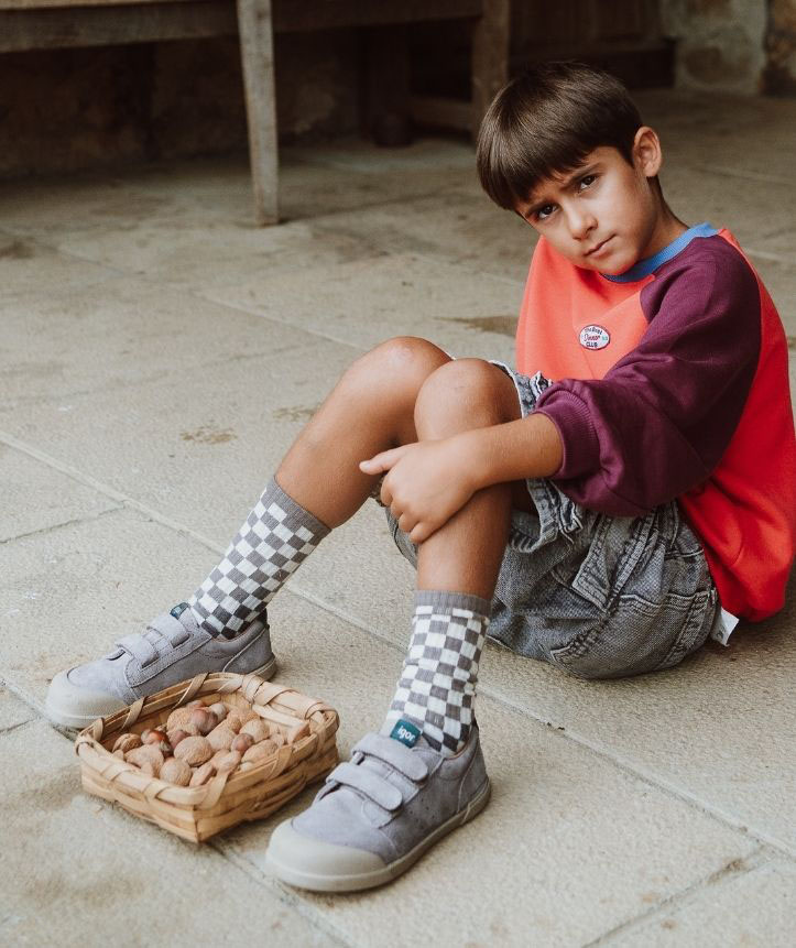 Criança com meias quadriculadas, sapatos cinza e camisola vermelha sentada ao lado de uma cesta de nozes